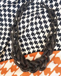 A necklace with that has a thick, gunmetal, woven chain necklace. It is on top of a black, orange, and white hounds-tooth scarf 