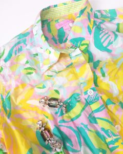 Close up of the top portion of a Lilly Pulitzer Hawaiian style shirt. The shirt is colored in bright yellow, green, aquas, and pinks and has a pair of crystal like dangle earrings on top of it.