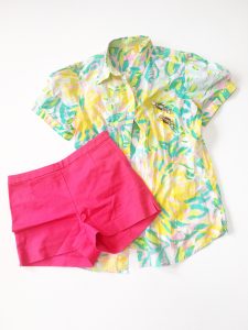 A Lilly Pulitzer Hawaiian style shirt. The shirt is colored in bright yellow, green, aquas, and pinks. With the shorts is a pair of bright pink shorts