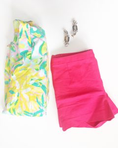 A Lilly Pulitzer Hawaiian style shirt. The shirt is colored in bright yellow, green, aquas, and pinks. With the shorts is a pair of bright pink shorts. The shirt and shorts are folded up. There is also a pair of crystal like dangle earrings