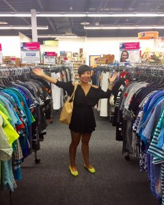 The DCGF (a woman with short, black hair in a short black dress and neon yellow shoes, stands between two racks of clothing in the Goodwill of Greater Washington Sully Station store