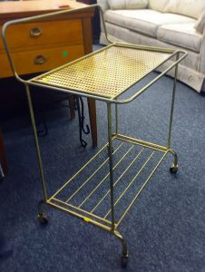 A gold bar cart made of brass. It has two levels and has black swivel wheels 