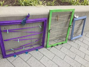 A picture of three frames with the glass and backing taken out and replaced with a zig zag ribbon to hang items on. There is a purple one with a bird sitting on top, a green one in the middle, and a third light blue one on the right with a small white bird on it.