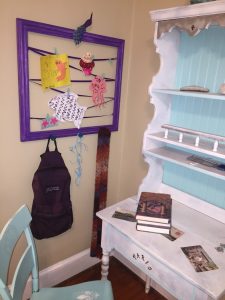 A picture of a purple frame with a zig zag purple ribbon attached to it that holds different cards and children's projects and it has a backpack hanging from the bottom. Next to it is a white desk and a blue chair