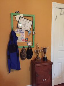 A green frame with a zig zag twine on it that holds certificates and pictures and there is a coat and a couple of hats handing from the bottom of it. There is a small side table next to it that holds two trophies