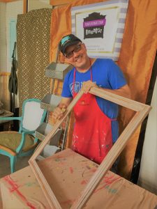 Tim Kime (a man with glasses in a red smock, blue shirt, and black cap) sanding a large picture frame