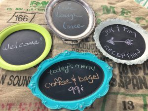 A picture of four silver serving trays that have been painted with colorful paint around the edges and then with chalk board paint in the center so messages can be written on them. There is a vertical oval one at the top that says live, laugh, love. A lime green one on the left that says welcome, a light blue one on the bottom that says today's menu coffee and bagel $.99, and a white one on the right that says Joy and Tim Reception with an arrow pointing toward the left.