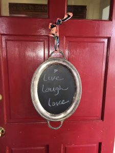 A silver serving tray that has been painted in the center with black chalkboard paint with the words live laugh love written in blue chalk on it. It is hanging by a colorful ribbon on a red door