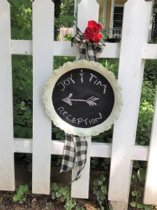 A silver serving tray that has been painted with a white boarder and black chalkboard paint in the middle with Joy and Tim Reception with an arrow drawn on it in pink chalk. It is hanging from a white picket fence with a white and black checkered ribbon and a red rose at the top