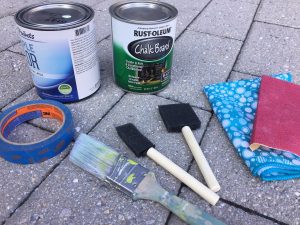 A picture of the materials needed to DIY the silver trays: a couple of cans of various latex and chalkboard paints, a roll of blue painters tape, three different brushes (one regular and two sponge), a blue rag, and sand paper
