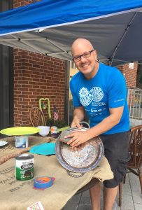 A picture of Tim Kime (a man in a blue t-shirt, bald, with glasses and wearing cargo shorts) working on one of the DIY silver serving trays