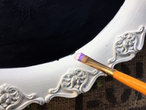 A picture of a silver serving tray that has been painted with a white boarder and a black chalk board paint inside. You can see the tip of a small brush with white paint on the tip.