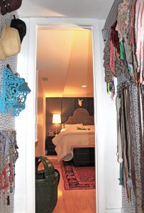 A picture from the inside of the closet out into the bedroom. In the bedroom there is a bed with a white comforter, a large headboard, a red carpet, a cow skill above the bed, wooden floors, and a side table with a lamp