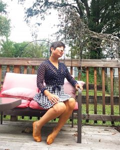 A picture of a woman with short dark hair sitting outside on a patio on a seat with pink coushions. She is wearing a sheer long-sleeved top, colored with navy blue and taupe polka dots, by Italian high-end designer, Fuzzi and a vintage houndstooth black and white skirt,