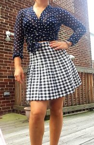 A picture of a woman from the neck down. She is wearing a sheer long-sleeved top, colored with navy blue and taupe polka dots, by Italian high-end designer, Fuzzi and a vintage houndstooth black and white skirt, 