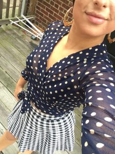 A woman taking a selfie. It is pointed down so you cannot see her face. She is wearing a sheer long-sleeved top, colored with navy blue and taupe polka dots, by Italian high-end designer, Fuzzi and a vintage houndstooth black and white skirt, and a pair of gold, bone, hoop earrings