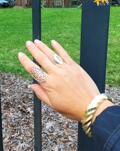 A picture of the back of the DC Goodwill Fashionista's hand. She is wearing a copper ring that covers most of her pointer finger that has designs cut into it. She is also wearing a ring on her ring finger that has an oval shape and has a slit cut into it. She is wearing a gold twist bracelet on her wrist. In the background is a black fence and the ground has mulch on it an there is green grass.