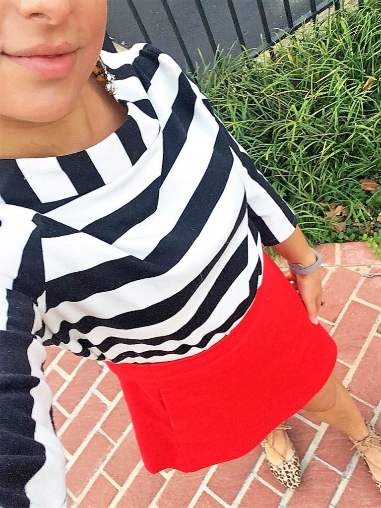 A picture of the DC Goodwill Fashionista. She is taking a top down selfie and you can see her mouth and chin and the whole front of her body. She is wearing a black and white striped boatneck shirt, a red skirt, and a pair of leopard print shoes. She is standing on a brick sidewalk with some grass and a fence in the background