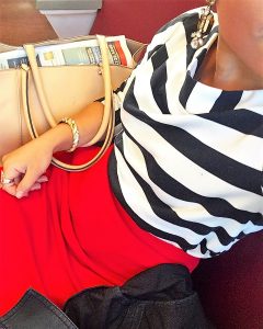 A picture of the DC Goodwill Fashionista. She is taking a top down selfie and you can see her mouth and chin and the whole front of her body. She is wearing a black and white striped boatneck shirt, a red skirt, a gold twisted bracelet, and has a tan purse and dark coat next to her. She is sitting on the Metro train