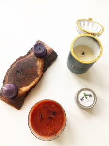 A top down view of five candles. One with Two small purple tea candles on either end of a larger wooden natural looking candle holder. Another of a ceramic cylinder containing a candle. The ceramic is white and has a blue, gold, and black chevron pattern on it. In the center there is a black label with the words "Maison" in gold and "Sandalwood" in blue. Another of an orange candle in a glass, cylinder container. On the front there is a label saying Yankee Candle with a picture of pumpkins and the words "Spiced Pumpkin" on it. And the last and smallest one is of a small cylinder shaped tin (shown from the top down). On the top it has a white label with two black elephants with a green tree in between them. The words "One Kings Lane" is written in all capital letters and the website www.allkingslane,com is under the capitalized words 