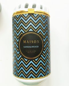 A picture of a ceramic cylinder containing a candle. The ceramic is white and has a blue, gold, and black chevron pattern on it. In the center there is a black label with the words "Maison" in gold and "Sandalwood" in blue 