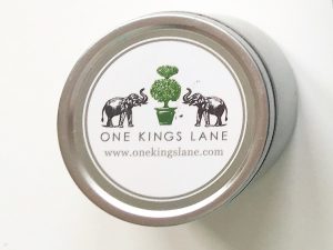 A picture of a small cylinder shaped tin (shown from the top down). On the top it has a white label with two black elephants with a green tree in between them. The words "One Kings Lane" is written in all capital letters and the website www.allkingslane,com is under the capitalized words 