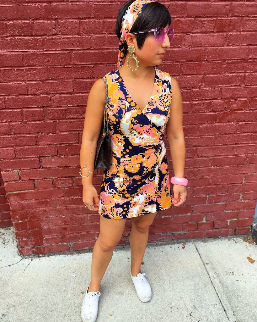 A picture of a woman in front of a red painted brick wall. The woman is the DC Goodwill Fashionista. She has short, brown hair and is wearing a 60's. mod, patterned, mini dress. The dress is yellow, purple, pink, and white and she has a headband that has the same pattern and colors. She is wearing white tennis shoes, pink sunglasses, an assortment of gold jewelry, and has a brown purse.