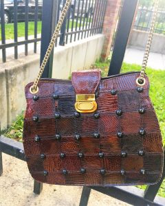 A picture of a brown purse with a gold chain hanging from a black metal fence. It is made out of brown, leather squares and has black studs and a gold clasp.