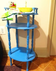 A blue side table with three layers with a yellow bowl, a colorful glass cup, and colorful cloth on top of it