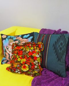 A picture of three pillows on a yellow couch with a purple blanket on it. In the center there is a brown pillow with a red and yellow floral pattern. On the right there is a pillow with a native american like pattern on it that is a dark shade of green with some brown, black, and red stripes. And on the left there is a pillow that is black but has blue and light brown paint stroke like patterns on it. 