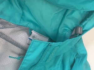 A picture of a blueish green, Arcadia-style, Columbia raincoat. It is a close up of the hood, tag, inner gray mesh, and the drawstring for the hood