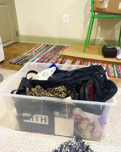 A large, plastic, storage bin that has a variety of items in it like clothing and bags. The bin looks like it has been used for moving. It is on top of a white carpet and there is a multicolor carpet in the background and a green chair.