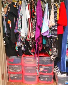 A picture of a closet. There is a large number of hanging clothes and below them are nine stackable plastic pink drawers. You can see inside the drawers and there are various clothing items in them.