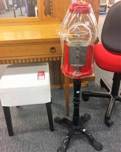 A vintage, red, gumball machine standing among a brown, wooden desk, a white footstool, and a red, rolling, office chair 