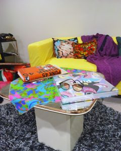 A side table in front of a yellow couch on top of a black, white, and gray shag carpet. It has various colorful books and a cloth on top as well as a pair of glasses. The table itself has a retro style to it and has a white bottom and brown top