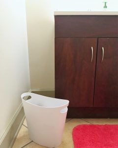 A small white trash can in a bathroom. You can see the brown sink cabinet and pink bath mat 