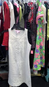 A picture of a rack of clothing in a Goodwill retail store. A white, sleeveless dress is not hanging with the rest of the clothes and is shown more prominent in the picture 