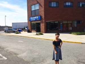 A picture of the DC Goodwill Fashionista (a woman in a knee length skirt and a black top with short black hair) standing in the parking lot outside of the Goodwill store on South Dakota Ave. in Washington DC