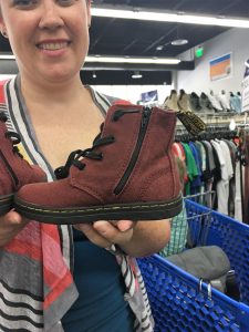 A woman in a Goodwill store holding a burgundy colored shoe. There are clothing racks and a blue shopping cart is in the background