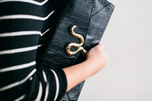 A picture of a woman's arm holding a black, leather, faux alligator skin, clutch purse with a gold snake ornamenting it.