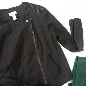 A close up of the black moto jacket. The zipper and button are clearer as well as the tag that says LOFT. The corner of the hunter green pants are also seen