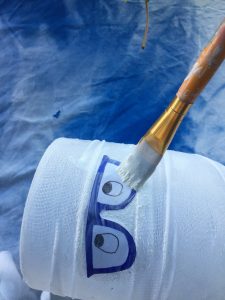 A picture of a set of cut out paper eyes with blue classes being adhered to a glass vase that has been wrapped in gauze. It is being adhered by a small paint brush using mod podge.