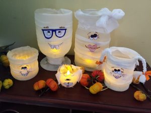 A picture of five glass containers wrapped to look like a mummy family as Halloween decorations. There is a father with blue glasses, a mother with a bow, a daughter with pig tails, a son with large front teeth, and a dog with floppy ears. They have been made wrapped with gauze 