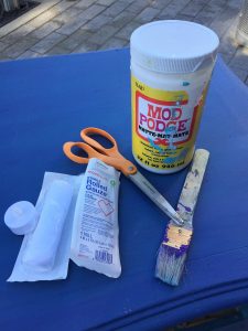 A picture of the various materials needed for the project. These include medical gauze, scissors, a paint brush, and mod podge 