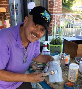 A picture of Tim Kime (a man wearing a black baseball cap and a purple shirt with a goatee working on one of the DIY mummies. He is smiling at the camera