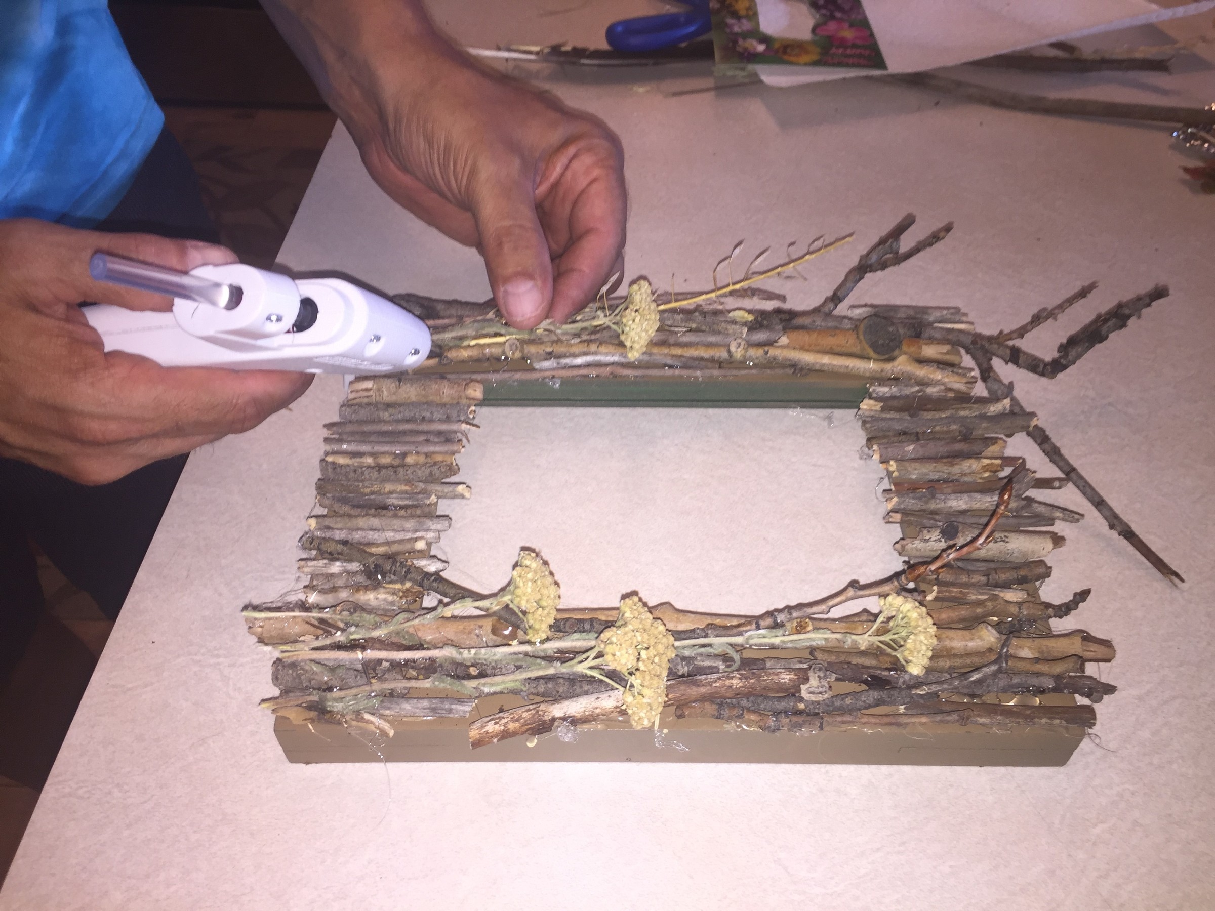 Tim hot glues twigs and flowers to a painted picture frame.